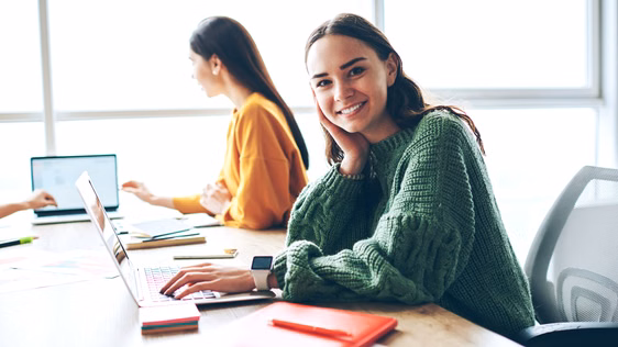 Porträt einer fröhlichen kaukasischen Frau, die auf dem Universitätsgelände am Laptop lernt; kompetente, hübsche Kolleginnen, die in einem Coworking Space arbeiten, eine WLAN-Verbindung nutzen und in die Kamera blicken