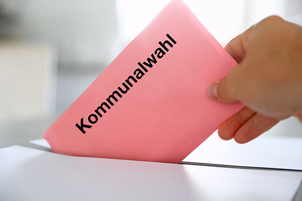Eine Hand, die einen rosafarbenen Umschlag mit dem Text Kommunalwahl in eine Wahlurne steckt