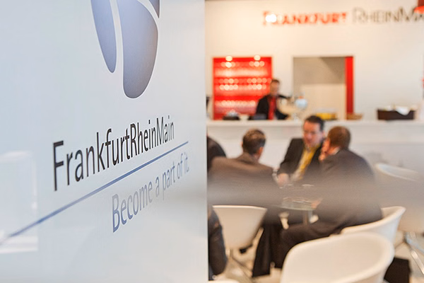 Im Vordergrund Logo FrankfurtRheinMain im Hintergrund Personen am Tisch und eine weitere am Empfang