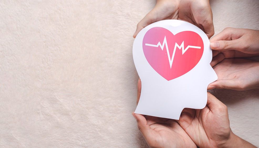 Hände halten eine weiße Papierfigur in Form eines Kopfes mit einem pinken Herzen und stilisiertem EKG-Linienverlauf auf dem Kopf