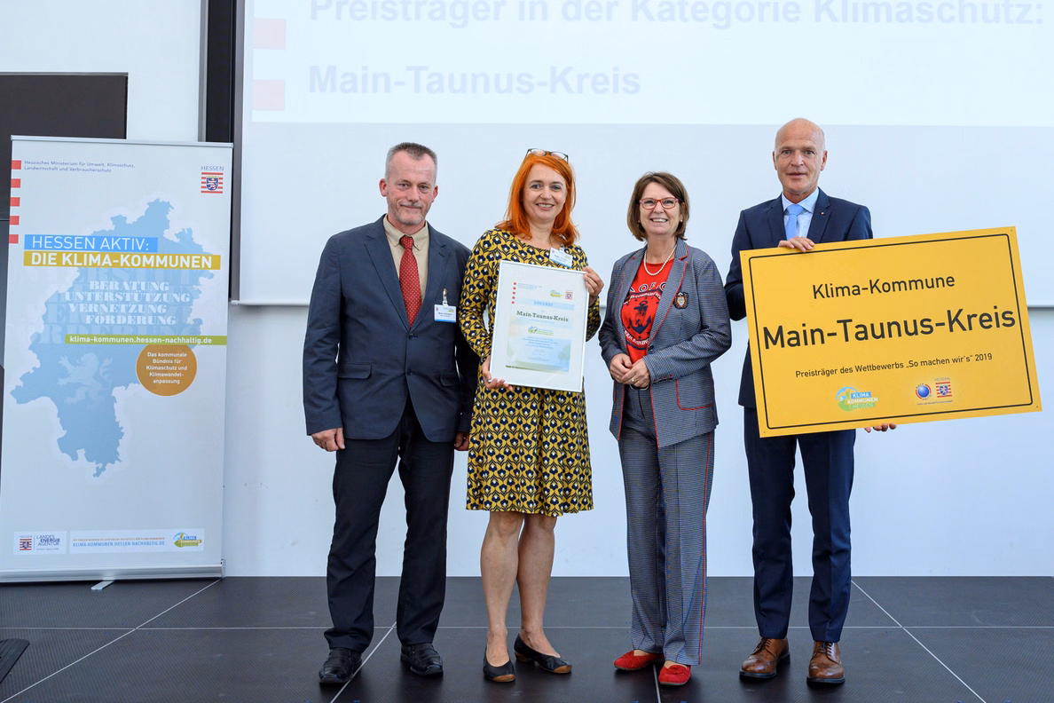 Zwei Frauen und zwei Männer, eine hält eine Urkunde in der Hand, ein zweiter ein großes Ortsschild mit dem Text Main-Taunus-Kreis