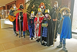 fünf Kinder in Verkleidung als Heilige Drei Könige, zwei Frauen und ein Mann vor einem geschmückten Tannenbaum