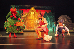 drei bunt verkleidete Personen auf einer Bühne, als Bühnenbild im Hintergrund sind eine Hütte, Bäume und ein Mühlrad zu sehen