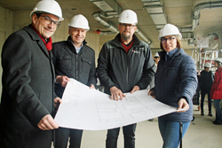 Bürgermeister Blasch, Landrat Cyriax, Peter Wesp (Leiter des Hochbau- und Liegenschaftsamts) und Schulleiterin Verena Rautenberg mit weißen Bauhelmen, die in einem Rohbau einen Bauplan in die Kamera halten