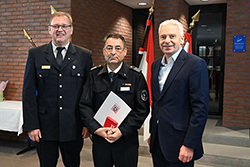 drei Männer in Anzug und Feuerwehruniformen, der Mann in der Mitte hält eine Urkunde