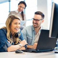 zwei Frauen und ein Mann, die lachend um einen PC-Arbeitsplatz sitzen und stehen