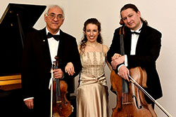 Ein Foto eines Ensembles in Trioformation, bestehend aus zwei Männern mit Violine und Cello und einer Frau. Im Hintergrund ist ein Klavier zu sehen.