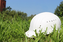 einen weißen Bauarbeiterhelm, der im Gras liegt. Im Hintergrund erscheint verschwommen das Eck eines rotbraunes Gebäude.