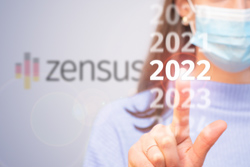 eine Grafik mit dem Text zensus 2022, im Hintergrund eine Frau mit Mundschutz, die mit dem Finger auf die Zahl 2022 tippt
