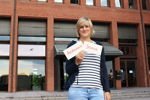 eine junge Frau vor dem Landratsamt. Sie hält zwei Zettel hoch, auf denen Ausbildung und Studium steht.