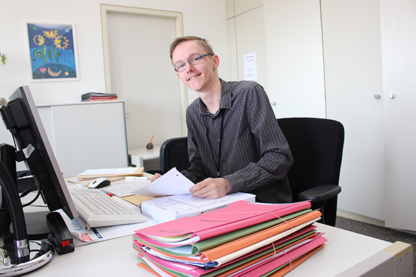 einen lächelnden jungen Mann an einem Schreibtisch mit Akten. 