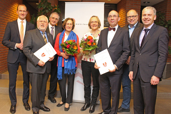Sechs Männer in dunklen Anzügen, zwei davon halten Urkunden in der Hand, und zwei Frauen mit Blumensträußen.