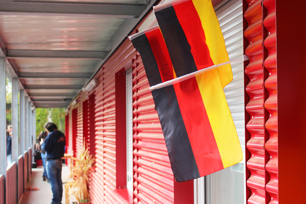 Einen schmalen Gang vor einem roten Wellblech-Gebäude, an dem Deutschland-Fahnen hängen.