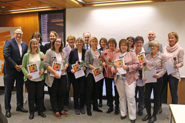 Eine Gruppe von etwa 15 Frauen und drei Männern, die in einem Raum vor einer weißen Wand stehen und in die Kamera lächeln.