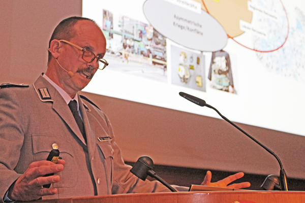 einen Mann mit einem Rednerpult, auf dem ein Mikrofon steht; im Hintergrund ein Teil einer Präsentation an der Wand.