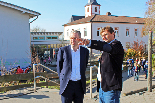zwei Männer in einem Innenhof zwischen verschiedenen Gebäuden, die auf etwas zeigen - im Hintergrund einige Kinder und eine Kirche.