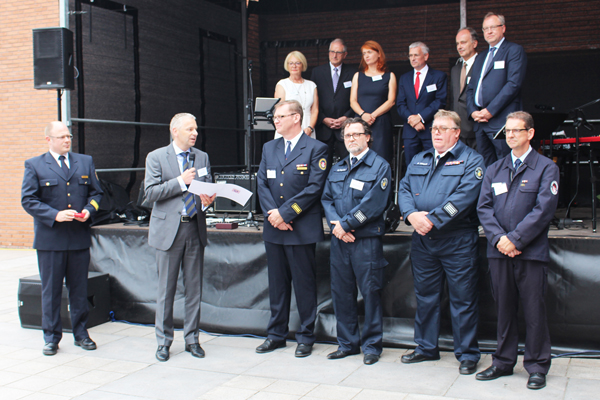 eine Gruppe Männer in Feuerwehr-Uniformen vor einer Bühne und einige Personen auf der Bühne; im Vordergrund ein Redner im Anzug.