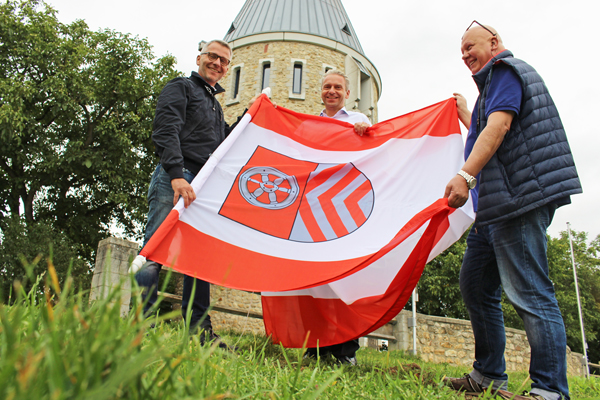 drei Männer vor einem Turm mit einer großen Fahne mit MTK-Wappen in den Händen