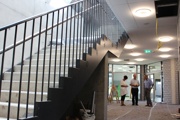 ein Gebäude im Rohbau mit einer Treppe im Vordergrund und drei Personen, die sich im Hintergrund unterhalten