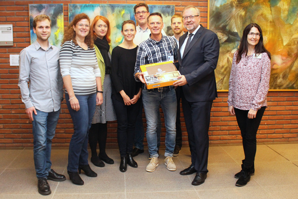 eine Gruppe Personen vor einer Wand, an der Bilder hängen; ein Mann übergibt einem anderen einen Preis