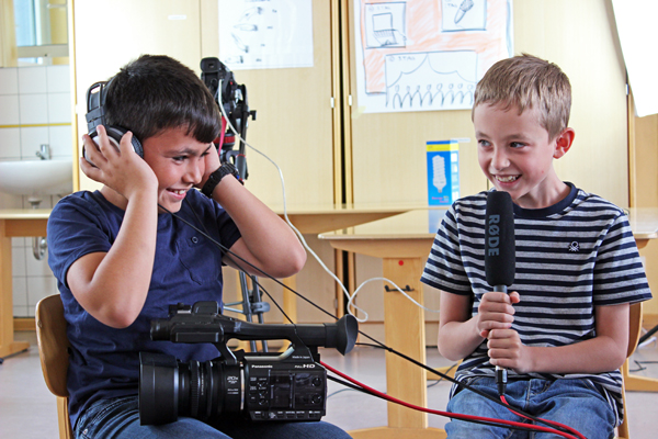 zwei Jungen, die mit einer Videokamera, Mikrofon und Kopfhörern lachend in einem Klassenraum sitzen