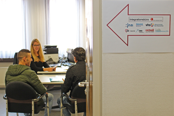 den Blick in ein Büro, in dem drei Personen sitzen; neben der Tür hängt ein Schild Integrationsbüro