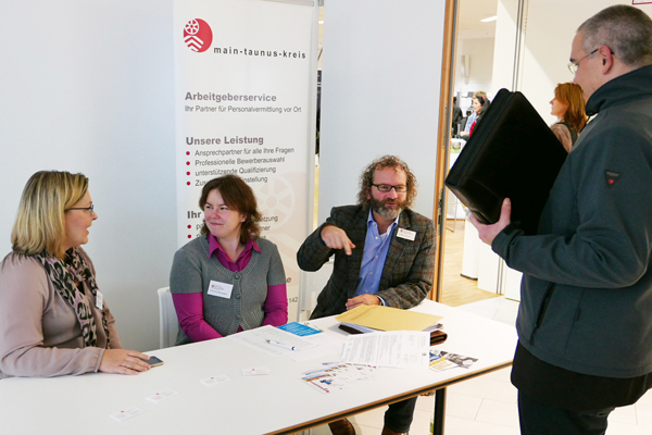 drei Personen an einem Tisch sitzend, davor steht ein weiterer Mann; im Hintergrund ein Plakat des Main-Taunus-Kreises zum Arbeitgeberservice