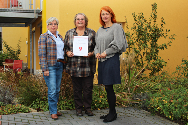drei Frauen, die mittlere mit einer Urkunde in der Hand, vor einem gelb gestrichenen Gebäude