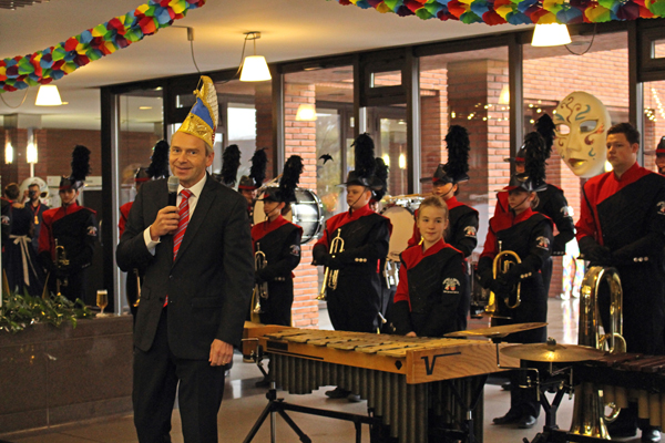 einen Mann im Anzug mit Narrenkappe und Mikrofon in einem bunt geschmückten Raum; im Hintergrund eine Musikkapelle