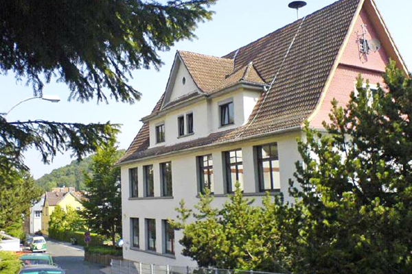 ein älteres Haus mit steilem Dach, das teilweise von Büschen und Bäumen verdeckt wird