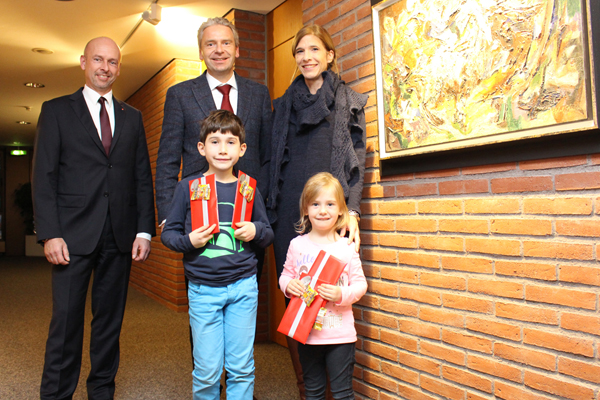 zwei Kinder mit Geschenken in der Hand und drei erwachsene Personen in einem Gang neben einem großen Bild