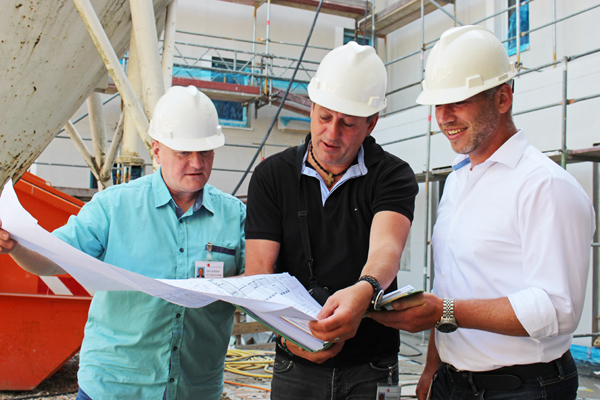 drei Männer mit weißen Bauhelmen, die auf einer Baustelle stehen und einen großen Bauplan in den Händen halten