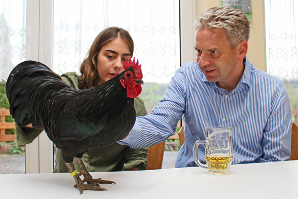 im Vordergrund ein schwarzer Hahn auf einem Tisch, der von einem Mädchen und einem Mann angeschaut wird