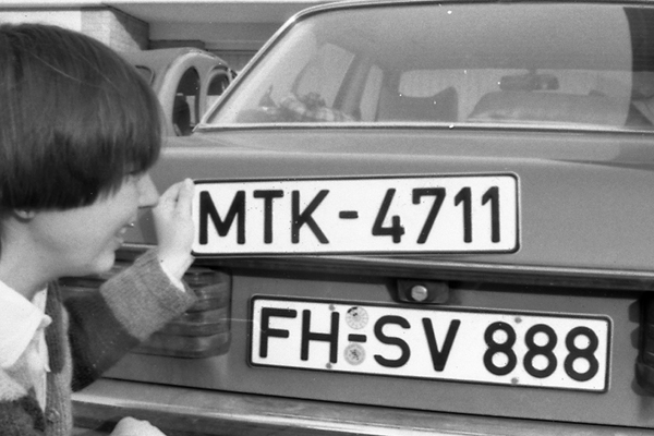 ein Schwarzweißbild von einer Person mit einem Nummernschild in der Hand, die neben dem Heck eins Autos kniet