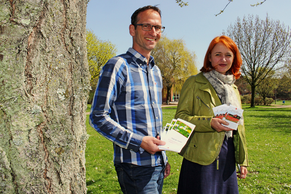 ein Mann und eine Frau mit Broschüren in der Hand, die auf einer Wiese neben dem Baum stehen