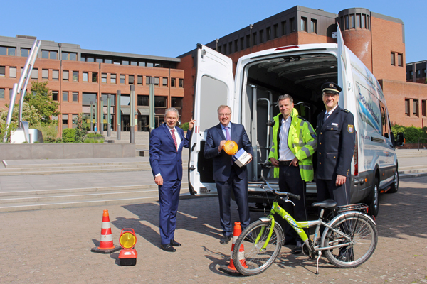 vier Männer in Anzügen und Uniformen vor einem Lieferwagen, vor dem ein Fahrrad, Pylonen und anderes Zubehör stehen
