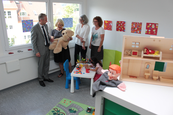 Ein Raum, weiß gestrichen, mit zwei Fenstern auf der linken Seite und Kinderspielzeug im Raum verteilt. Im Vordergrund rechts ein Puppenhaus und eine Handpuppe auf einer weißen Kommode. Im Hintergrund in der Mitte des Raumes vier Personen, eine davon hält