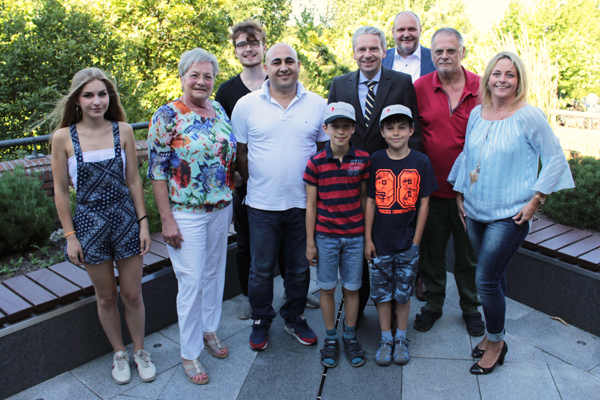 Zehn Menschen stehen in einer Gruppe auf einer Terrasse. Im Hintergrund sieht man grüne Bepflanzung. Die Gruppe schaut zur Kamera, im Vordergrund stehen zwei Kinder mit Schildmützen.