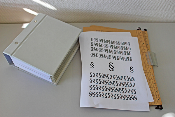 einige Ordner und Mappen auf einem Tisch, darauf ein Blatt Papier mit einigen Paragraphen-Zeichen