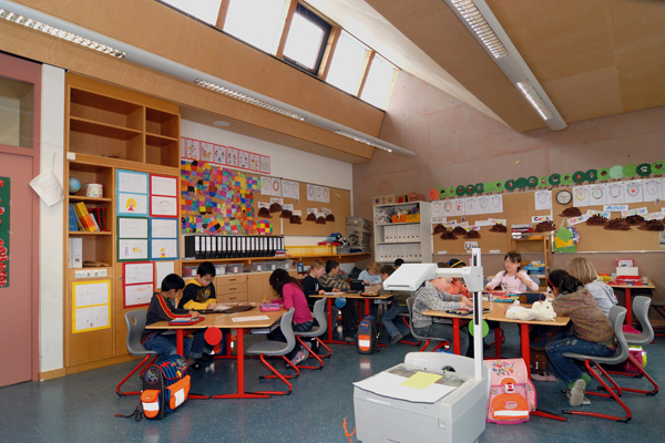 Einen Klassenraum mit Schülern, die an Tischen sitzen. Der Raum ist verziert mit Kinderzeichnungen auf Papier, es stehen viele Ordner im Hintergrund. Im Vordergrund sieht man einen Overhead-Projektor.