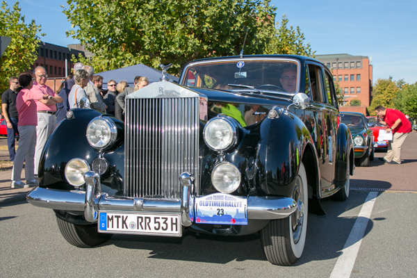 einen dunkelblauen Oldtimer von Rolls Royce, der über eine Straße fährt und von einigen Personen angeschaut wird