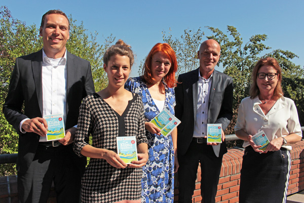 drei Frauen und zwei Männer, die kleine und bunte Bücher in der Hand halten, vor Büschen und blauem Himmel