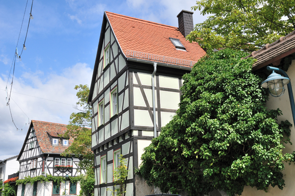 Zwei Fachwerkhäuser mit roten Dachziegeln und rechts viel Grünbewuchs. Im Hintergrund ein blauer Himmel mit vereinzelten Wolken.