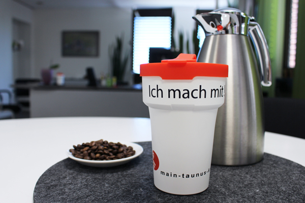 einen Kaffeebecher mit der Aufschrift Ich mach mit auf einem Tisch zusammen mit einer Kaffeekanne und einem Teller Kaffeebohnen