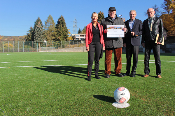 drei Männer und eine Frau auf einem Fußballplatz, im Vordergrund ein Fußball