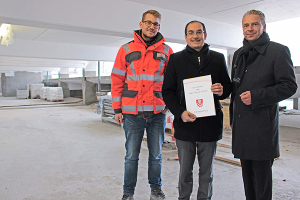 zwei Männer in dunklen Jacken und ein Mann in Warnjacke auf einer Baustelle; einer hält eine Urkunde in der Hand
