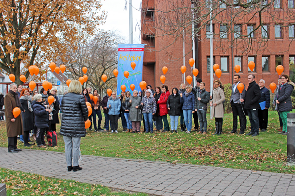 eine Gruppe Personen mit orangefarbenen Luftballons in den Händen vor einem Gebäude, an dem eine Fahne gehisst wird