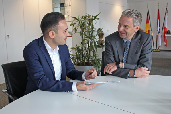 Zwei Personen unterhalten sich sitzend an einem Tisch in einem Büro. Die eine Person hält ein Tablet in der Hand; beide schauen sich an.