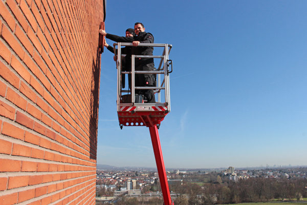 Zwei Männer stehen auf einer Hebelbühne und bringen an einer roten Steinwand einen Kasten an. Im Hintergrund sieht man blauen Himmel und einzelne Häuser