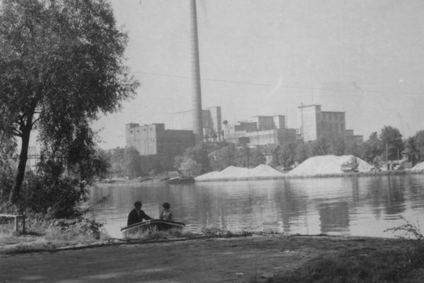 ein Schwarzweißfoto eines Ruderbootes mit zwei Personen an einem Flußufer; auf der anderen Flußseite eine Fabrik